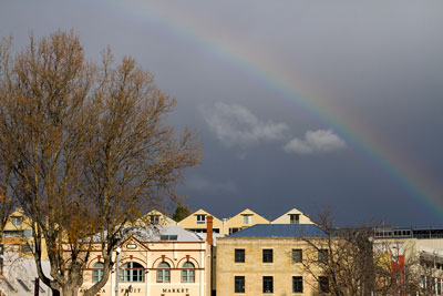 Houses in Hobart with the afternoon light blazing upon them, against a dark grey background. A rainbow cuts through the sky. Find out more things to do in Hobart.