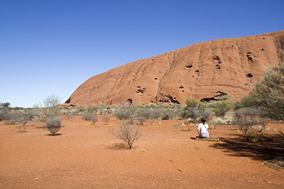 A woman sits on a log staring up at Uluru on a sunny, blue-skied day. Discover the best time to visit Uluru.