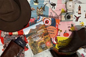 A collection of Australia themed icons, including an Akubra, earrings with an Aussie bird, a passport, Best Walks Australia and Welcome to Country books, RM Williams boots and a camera. Discover Australia travel essentials to pay for a trip to Oz.