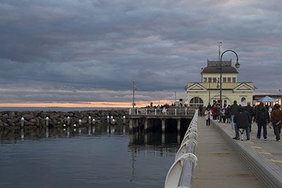 View of St Kilda pier at sunset in winter. People wander down the pier to the building at the end of the jetty. A visit to St Kilda is a must-do for any Melbourne itinerary.